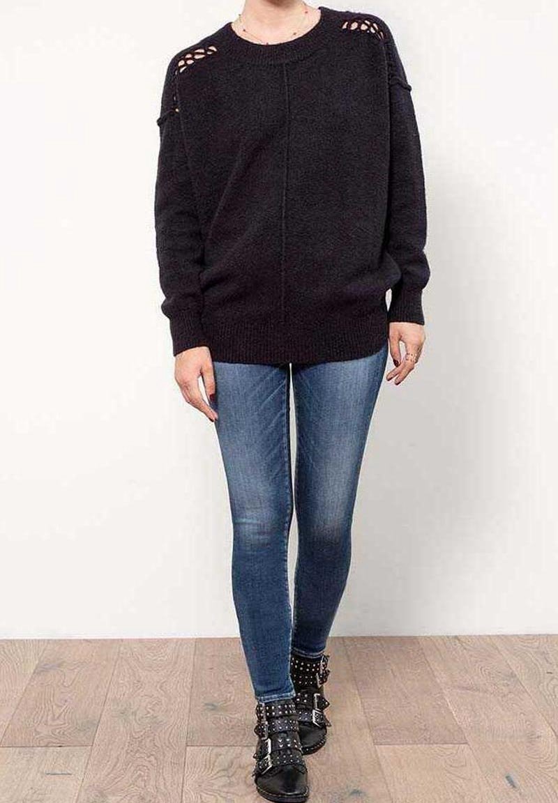 Personne portant un pull noir avec des découpes aux épaules, un jean skinny bleu et des bottines noires cloutées, debout sur un sol en bois.