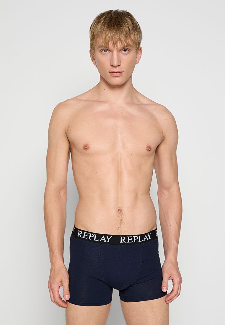 Jeune homme avec des cheveux blonds courts, portant des boxers bleu marine avec une ceinture noire marquée "REPLAY," se tenant contre un fond uni.
