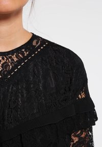 Top en dentelle noire avec un motif floral, encolure ronde avec bordure et accents froncés le long des manches, créant une texture en couches.