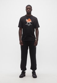 Άνδρας που φοράει μαύρο t-shirt Puma με πορτοκαλί γραφικό μπάσκετ και αντίστοιχο μαύρο παντελόνι και αθλητικά παπούτσια, στέκεται μπροστά σε λευκό φόντο.