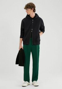 Felpa nera con zip abbinata a pantaloni sportivi verdi; modello che indossa sneaker bianche, mentre tiene una giacca nera texturizzata.