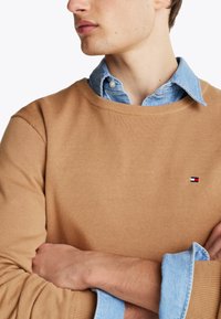Pulover tricotat de culoare bej, cu guler rotund, suprapus peste o cămașă din denim de culoare albastru deschis. Prezintă un mic logo roșu, alb și albastru pe piept.