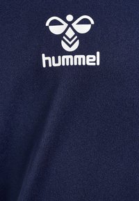 Białe logo Hummel ze stylizowaną pszczółką i tekstem na ciemnoniebieskim, teksturowanym materiale.