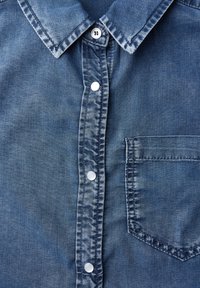 Dunkelblaue Jeanshemd mit Knopfleiste, spitzem Kragen und einer Brusttasche auf der linken Seite. Der Stoff hat eine weiche, strukturierte Oberfläche.