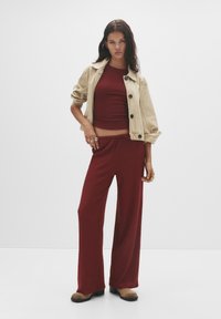Giacca di denim beige con bottoni neri sopra una canotta a coste bordeaux e pantaloni a gamba larga. Sottile texture con una vestibilità comoda. Calzature marroni.