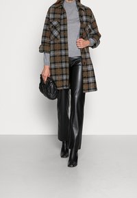 Femme portant un manteau à carreaux marron et gris par-dessus un col roulé gris, pantalon en cuir noir, bottes noires, tenant une pochette matelassée noire.