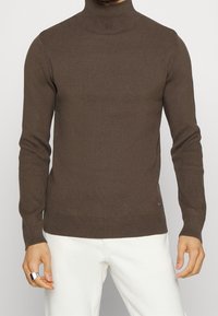 Pull à col roulé côtelé marron avec des manches longues, coupe ajustée et logo discret sur le côté inférieur, associé à un pantalon blanc.