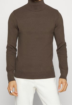 Brauner gerippter Rollkragenpullover mit langen Ärmeln, figurbetonter Passform und dezentem Branding an der Unterseite, kombiniert mit weißen Hosen.