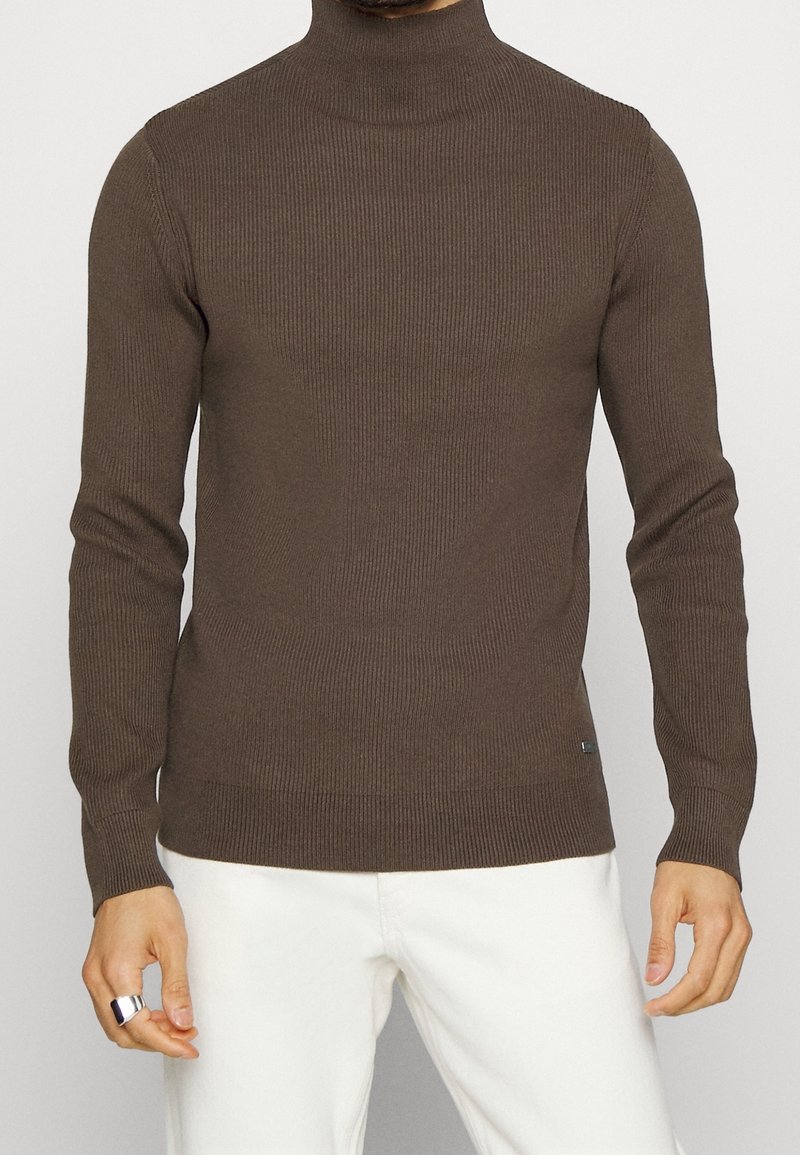 Pull à col roulé côtelé marron avec des manches longues, coupe ajustée et logo discret sur le côté inférieur, associé à un pantalon blanc.