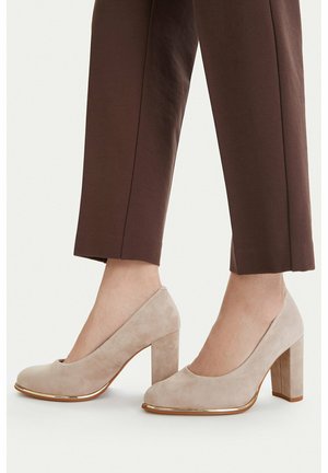 Zapatos de tacón grueso de ante beige con borde dorado, usados con pantalones marrones recortados, mostrando vistas laterales y frontales de ambos zapatos.