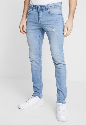 Vaqueros de mezclilla azul claro con un ajuste slim, que presentan detalles desgastados y un sutil desteñido, combinados con zapatos deportivos blancos.