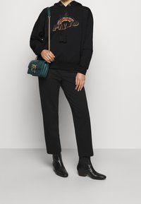 Haut à capuche noir avec un logo "PIMO" coloré brodé, associé à un pantalon noir et un sac à main matelassé vert. Des bottines noires complètent le look.