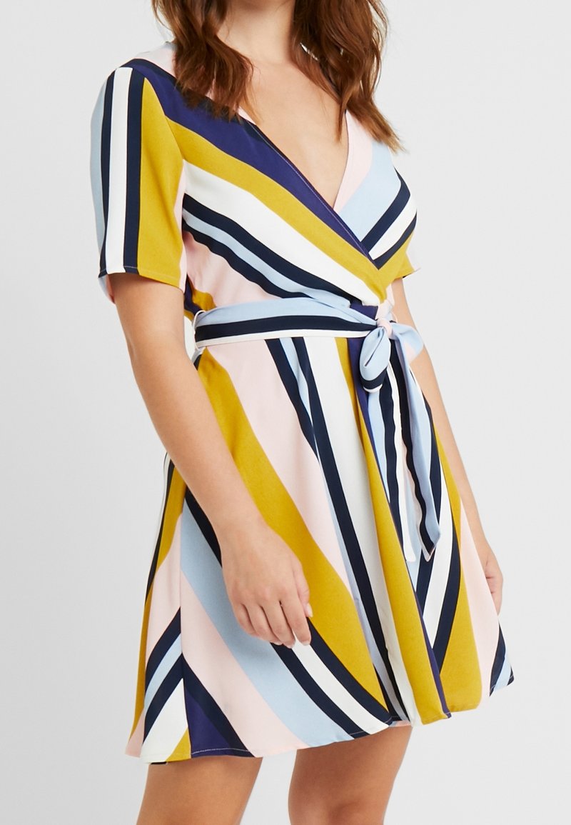Femme portant une robe portefeuille à manches courtes avec des rayures diagonales jaune, bleu marine, bleu clair, rose et blanc, nouée à la taille avec un nœud.