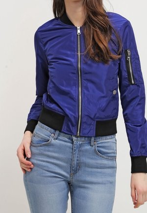 Kobieta w fioletowej kurtce bomberce zapinanej na zamek, z czarnymi mankietami i jasnoniebieskich dżinsach, stojąca na tle jednolitego tła.