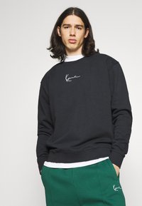 Hanorac negru cu un logo brodat alb pe piept, asortat cu pantaloni de trening verzi. Design casual cu guler rotund, mâneci lungi și manșete ribbate.