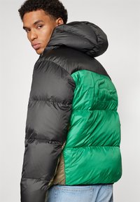 Chaqueta tipo plumas con una parte superior negra y una parte inferior verde, con una textura brillante y capucha, diseñada con acolchado en forma de rombos.