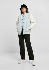 Svetlo modrá varsity bunda s bielymi rukávmi, pruhovanými manžetami a vyšitým logom. Skombinovaná s tmavými nohavicami a bielymi teniskami. Súčásťou je aj klobúk typu bucket.