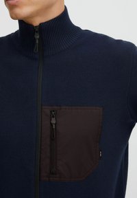 Sudadera de punto azul marino con cremallera y cuello alto, y un bolsillo con cremallera de color marrón oscuro en el lado izquierdo del pecho. Textura acanalada en el cuello y el cuerpo.