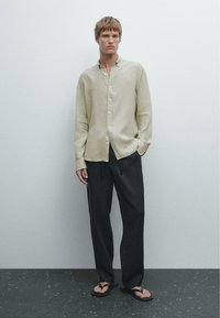 Beige linned skjorte med knapper og lige kant, parret med mørke, løstsiddende bukser og sorte flip-flops, med et minimalistisk design.