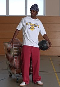 Osoba u bijelom majici kratkih rukava s logom Los Angeles Lakers drži košarkašku loptu, nosi crvene široke hlače i šešir s uzorkom.