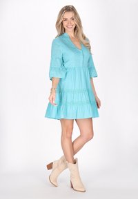 Rochie din bumbac turcoaz cu fusta în straturi, mâneci puff 3/4 și detalii cu gaură. Asortată cu bocanci cowboy bej până la gleznă.