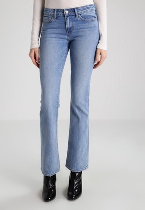 Jean droit bleu clair porté avec un haut blanc à manches longues et des bottines noires brillantes sur un fond neutre.