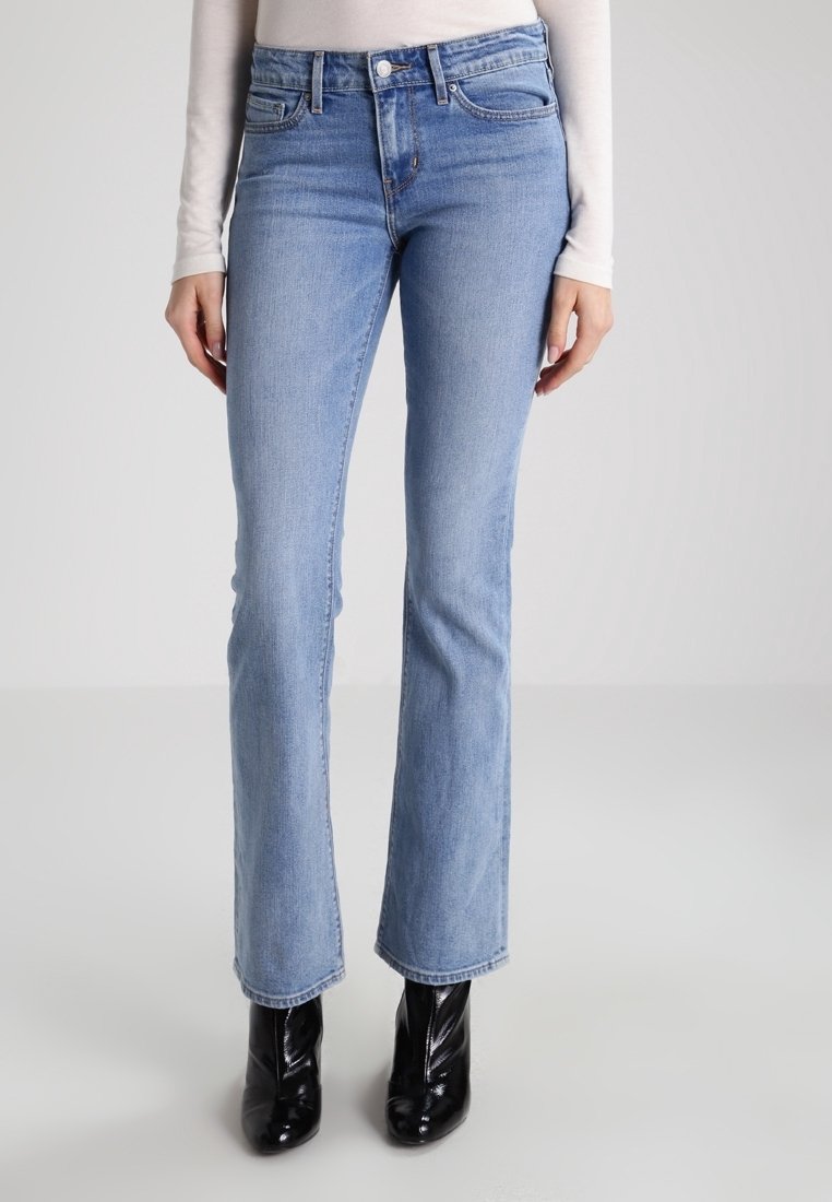 Jean droit bleu clair porté avec un haut blanc à manches longues et des bottines noires brillantes sur un fond neutre.