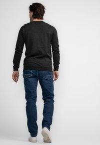 Zwarte trui met een ribbelboord ronde hals en manchetten, gecombineerd met blauwe jeans en witte sneakers, gezien vanaf de achterkant.