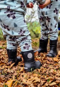 Svarta gummistövlar på barnens fötter, parat med ljusblå fleecepyjamasar med mönster av tvättbjörnar och träd, omgivna av fallna löv.