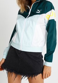 Chaqueta cortavientos Puma con cremallera frontal, diseño de bloques de color en azul claro, verde azulado y blanco, con acento de logotipo amarillo en la manga.