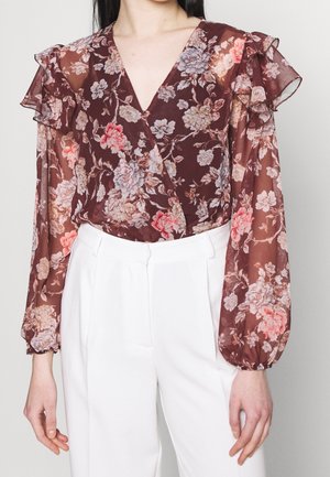 Vrouw draagt een doorschijnende bordeauxrode bloemenblouse met gerimpelde schouders en lange mouwen, gecombineerd met witte broek met een hoge taille.