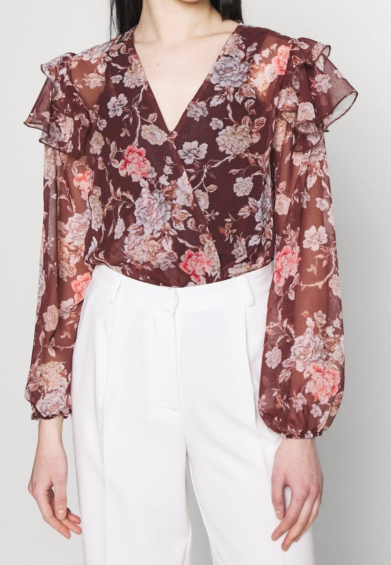 Femme portant un chemisier floral bordeaux transparent avec des épaules volantées et des manches longues, associé à un pantalon blanc taille haute.