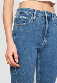 Jeans de mezclilla azul clásico con una cintura alta, diseño de cinco bolsillos, costuras visibles y parche con logo en el bolsillo trasero; textura suave.