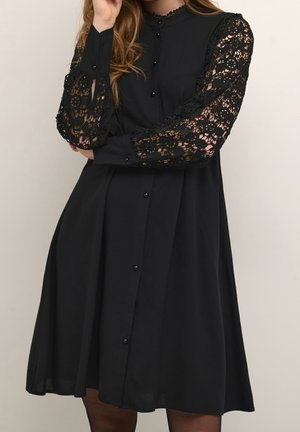 Woman wearing a black knee-length dress with a buttoned front and sheer lace floral sleeves, standing with one arm crossed and hand near face.