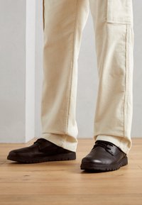 Chaussures en cuir marron à bout arrondi, surface texturée et semelle discrète ; associées à un pantalon en velours côtelé de couleur claire.