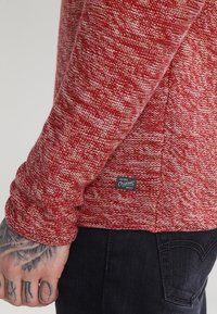 Röd texturerad långärmad skjorta med ett vävt mönster, utrustad med en etikett på den nedre sidan och svart denim synlig vid midjan.