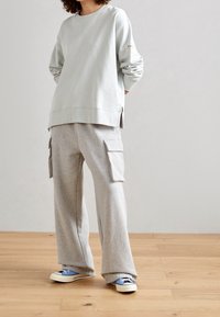 Světlý šedý svetr s žebrovanými manžetami a bočními rozparky, v kombinaci se šedými cargo kalhotami s bočními kapsami. Modro-bílé tenisky dotvářejí celkový vzhled.