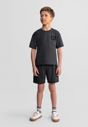 Chłopiec stojący, ubrany w czarną koszulkę z naszywką, czarne krótkie spodenki, paskowane skarpety i białe trampki na jednolitym jasnym tle.