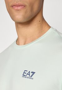 Svetlo mint zelena majica s kratkimi rokavi iz mehkega bombaža. Ima majhen temno moder logotip EA7 Emporio Armani na prsih. Gladka textura.