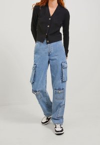 Pantaloni cargo in denim blu con multiple tasche laterali, vita alta, vestibilità rilassata e un lavaggio chiaro, abbinati a un cardigan nero con bottoni.