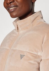 Giacca beige con zip caratterizzata da una morbida texture vellutata e colletto rialzato. Accentatura metallica a forma di triangolo sul petto.