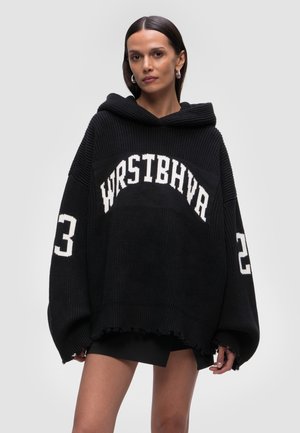 Femme portant un sweat à capuche noir tricoté surdimensionné avec capuche, texte blanc "WRSTBHVR" sur le devant et chiffres "23" sur les manches, debout devant un fond uni.