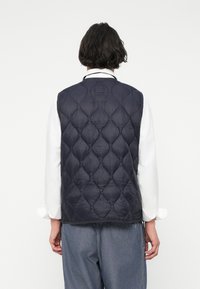 Ναυτικό γαλάζιο γαζωμένο γιλέκο με υφή διαμαντιού, που διαθέτει στρογγυλή λαιμόκοψη και χωρίς ορατούς κλειστές μηχανισμούς, φορεμένο πάνω από ένα λευκό πουκάμισο.