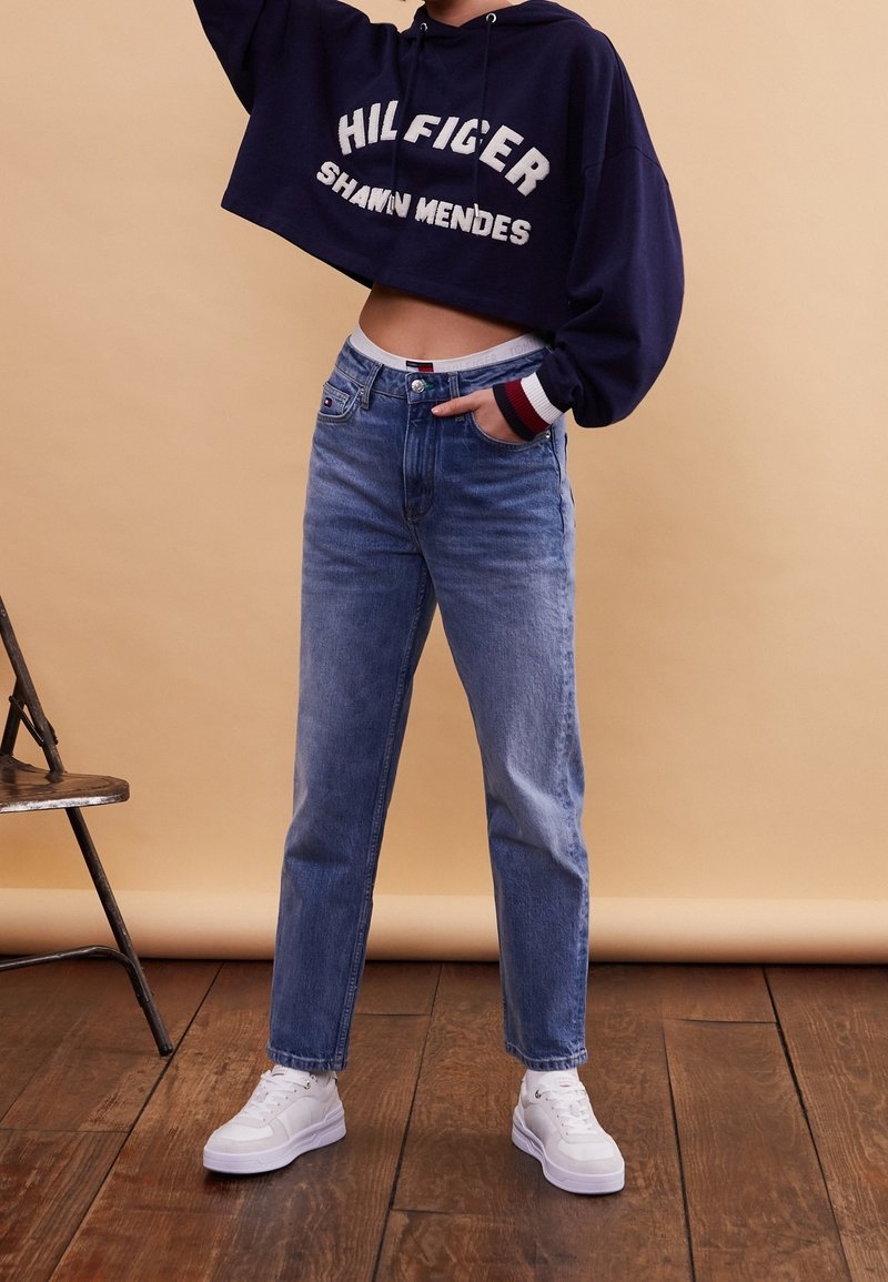Sweat à capuche court bleu marine avec logo blanc, jean taille haute bleu clair et baskets blanches, sur fond beige.
