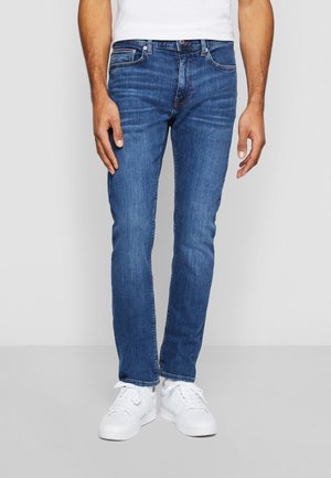 Jeans de mezclilla azul ajustados con un diseño clásico de cinco bolsillos, que presentan un acabado desgastado y sutiles detalles de costura, combinados con zapatillas blancas.