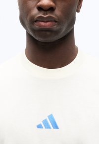 Κοντινό πλάνο ενός ατόμου που φοράει λευκό πουκάμισο με μικρό μπλε τριγωνικό λογότυπο στο στήθος, με απλό φόντο.