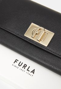 Juodas odinis piniginė su tekstūruota paviršiumi, auksiniu užsegimu su logotipu, ant baltos dėžutės su "Furla" prekės ženklu.