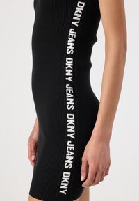 Μαύρο ριπ φόρεμα με λογότυπο DKNY Jeans σε λευκό κατά μήκος της πλευράς. Το ύφασμα φαίνεται ελαστικό και εφαρμοστό.