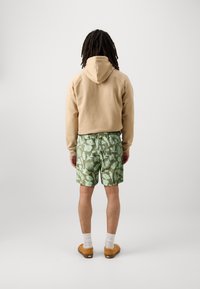 Sweat à capuche beige, short vert avec motif de feuilles, coupe décontractée, poches latérales. Sabots marron avec chaussettes blanches, se tenant sur un fond uni.