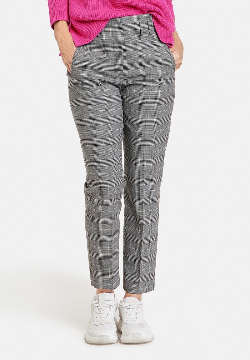 Personne portant un pantalon slim à carreaux gris, un pull rose vif et des baskets blanches, debout les mains dans les poches.